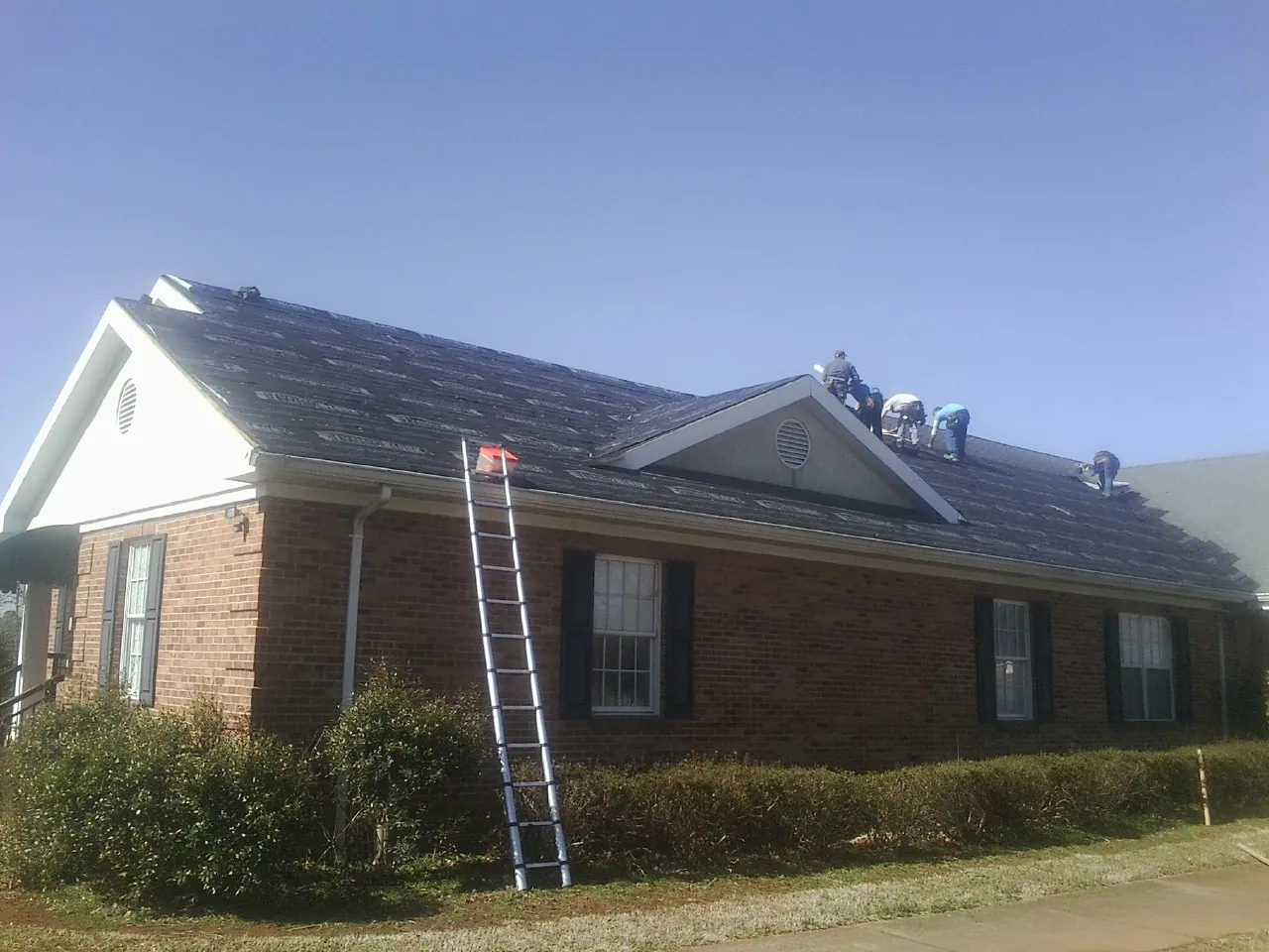 Crew tearing shingles off a brick ranch roof with a ladder set — Emergency Roof Repair work by our crew in Lee