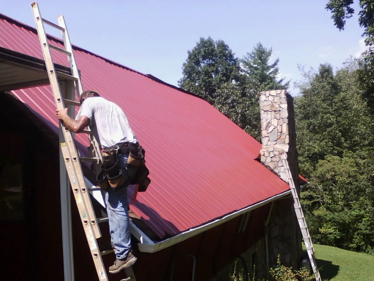 Roofer on a ladder beside a red metal roof and stone chimney — Soffit & Fascia Repair by Roofing Brothers in Lee